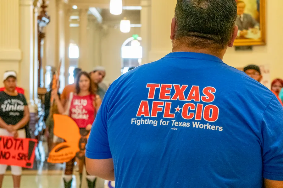 A man facing away from the camera wears a blue shirt with the text "TEXAS AFL-CIO" in red text. In the blurred distance, a crowd of activists stands in the rotunda of the Texas Capitol building.