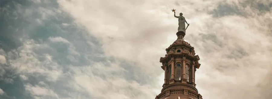 The top of the Texas State Capitol is captured before a cloudy sky.