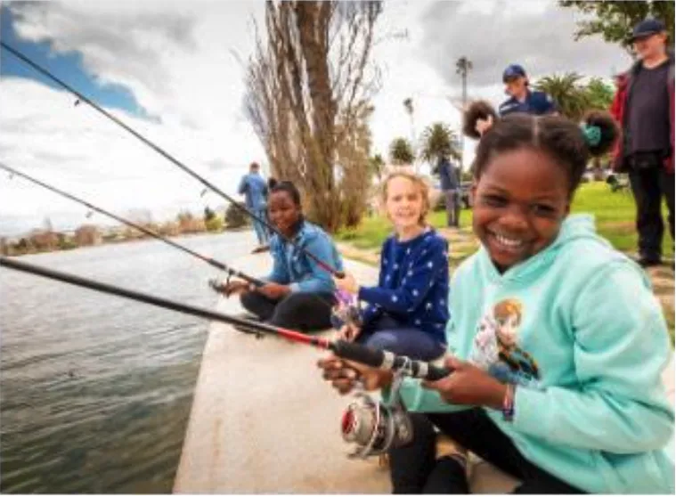 Three children sit along water's edge smiling and holding fishing poles.