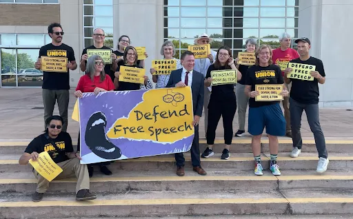Members of the Texas State Employees Union (TSEU-CWA Local 6186) stand in front of Hays County courthouse with signs that say "Defend Free Speech" in support of Dr. Tom Alter.